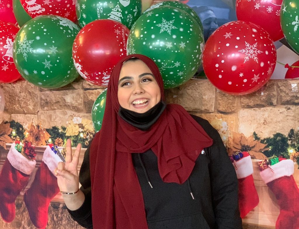 A female student wearing a black hoodie and red headscarf poses making a peace sign with her hands