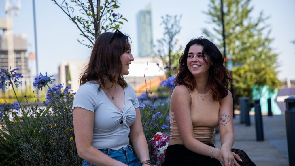 Two students sat together chatting. Manchester skyline in the background