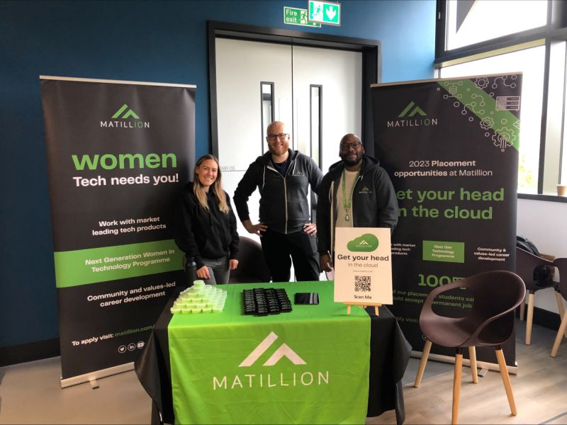 2 male and 1 female employees stand behind a table with a green matillion tablecloth and banners either side. The banner says ' women tech needs you!'