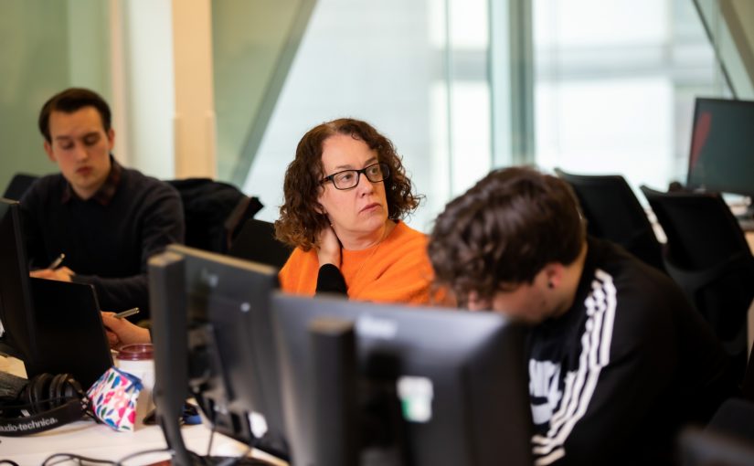 Erica in an orange jumper, sat in front of a computer beside course mates while listening to someone speaking.