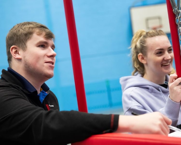 A young man with light brown hair, wearing a black Craghoppers jacket, sitting beside a young woman with blonde hair tied back, wearing a light purple hoodie, both smiling and engaging in conversation next to a red sports structure in an indoor sports facility with blue walls.