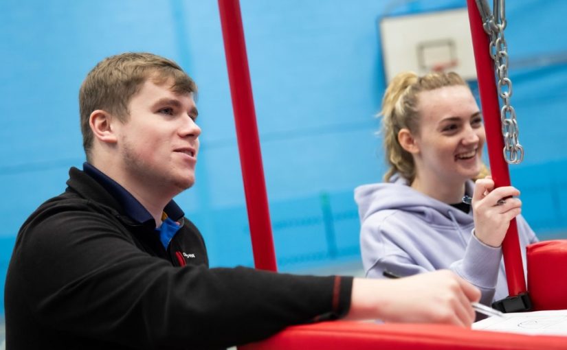 A young man with light brown hair, wearing a black Craghoppers jacket, sitting beside a young woman with blonde hair tied back, wearing a light purple hoodie, both smiling and engaging in conversation next to a red sports structure in an indoor sports facility with blue walls.