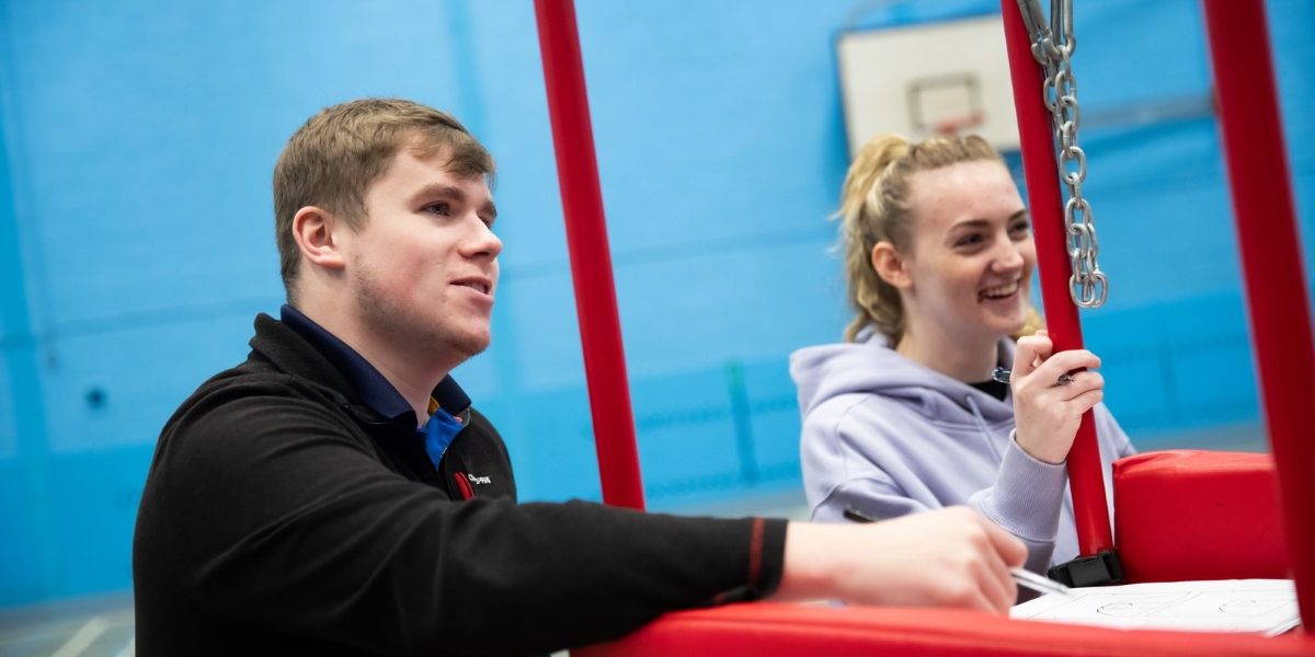 A young man with light brown hair, wearing a black Craghoppers jacket, sitting beside a young woman with blonde hair tied back, wearing a light purple hoodie, both smiling and engaging in conversation next to a red sports structure in an indoor sports facility with blue walls.